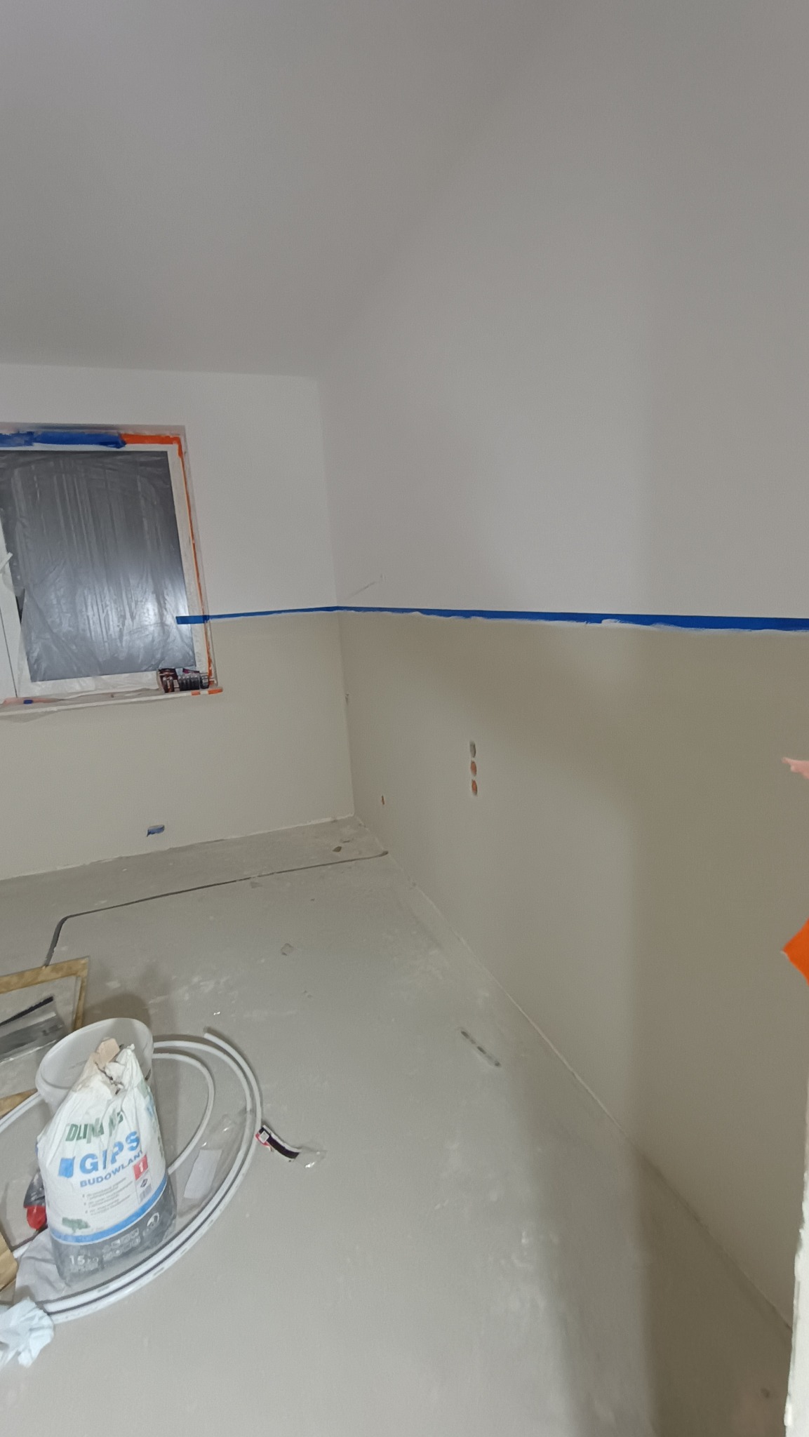 Pokój po szpachlowaniu, z widocznym workiem gipsu budowlanego, zabezpieczonym oknem i niebieską taśmą malarską na ścianach.