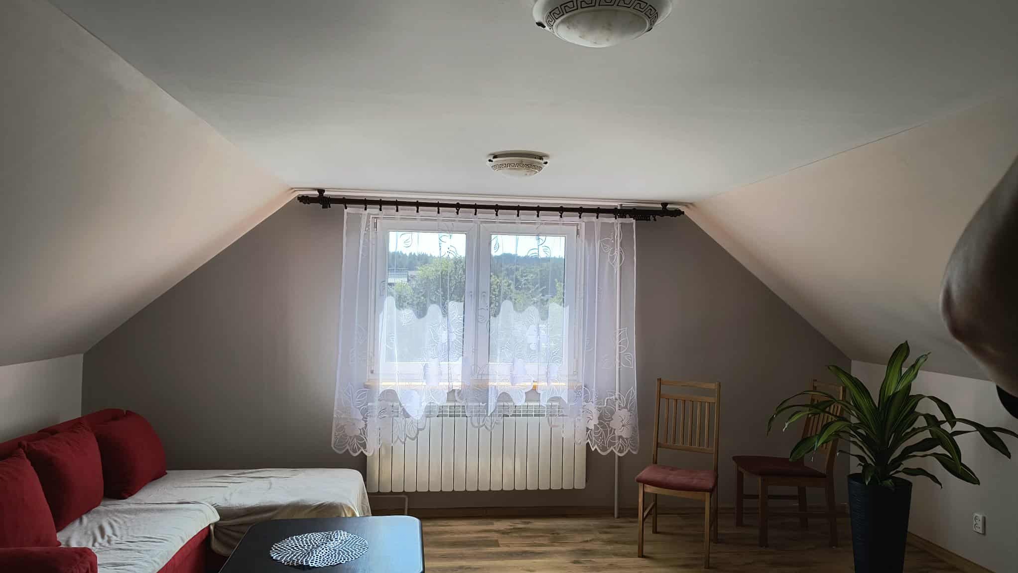 Pokój na poddaszu w Krakowie po odświeżeniu: szare ściany, drewniana podłoga, okno z firanką, czerwona sofa. Widoczne skosy i oświetlenie sufitowe.