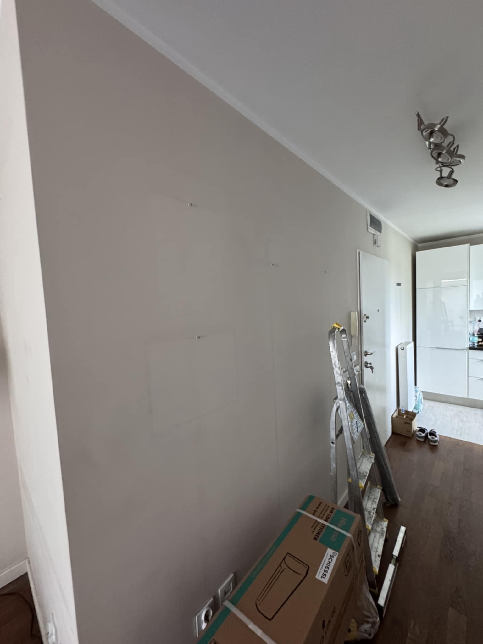 Ściana w mieszkaniu po szpachlowaniu, przygotowana do malowania. Widoczne ślady po szpachlowaniu, drabina i kartony w pomieszczeniu.