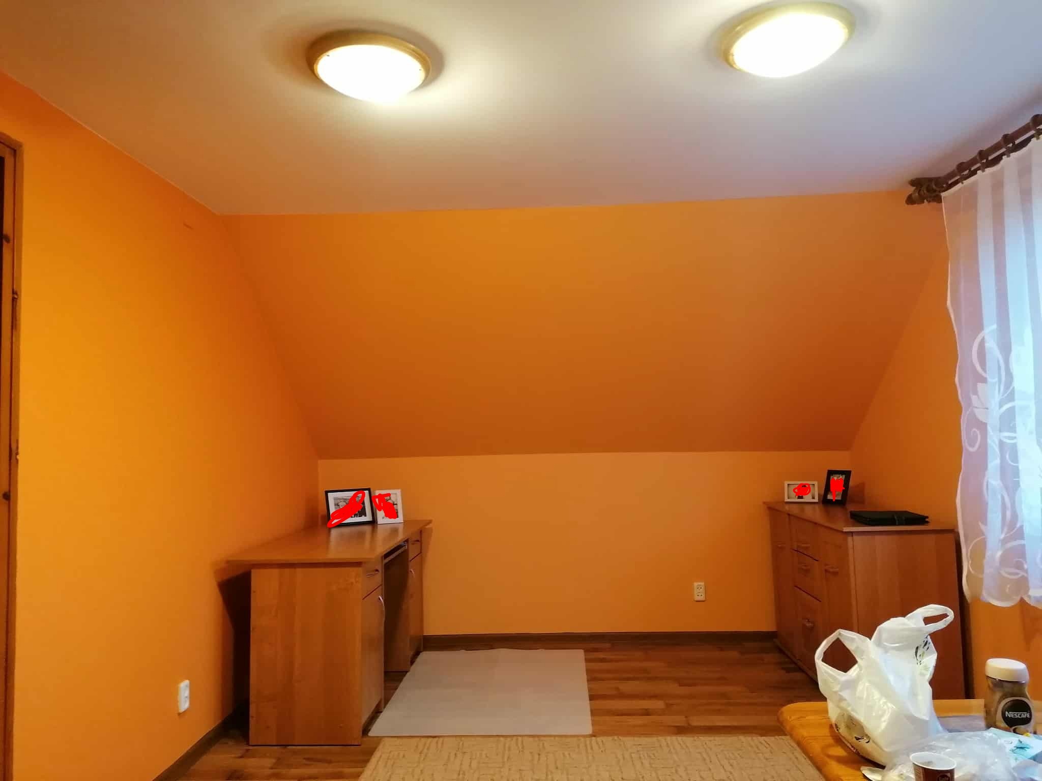 Pomarańczowe ściany na poddaszu po malowaniu, widoczne meble z jasnego drewna, okno z firanką i dywan na podłodze. Dwa punkty świetlne na suficie.