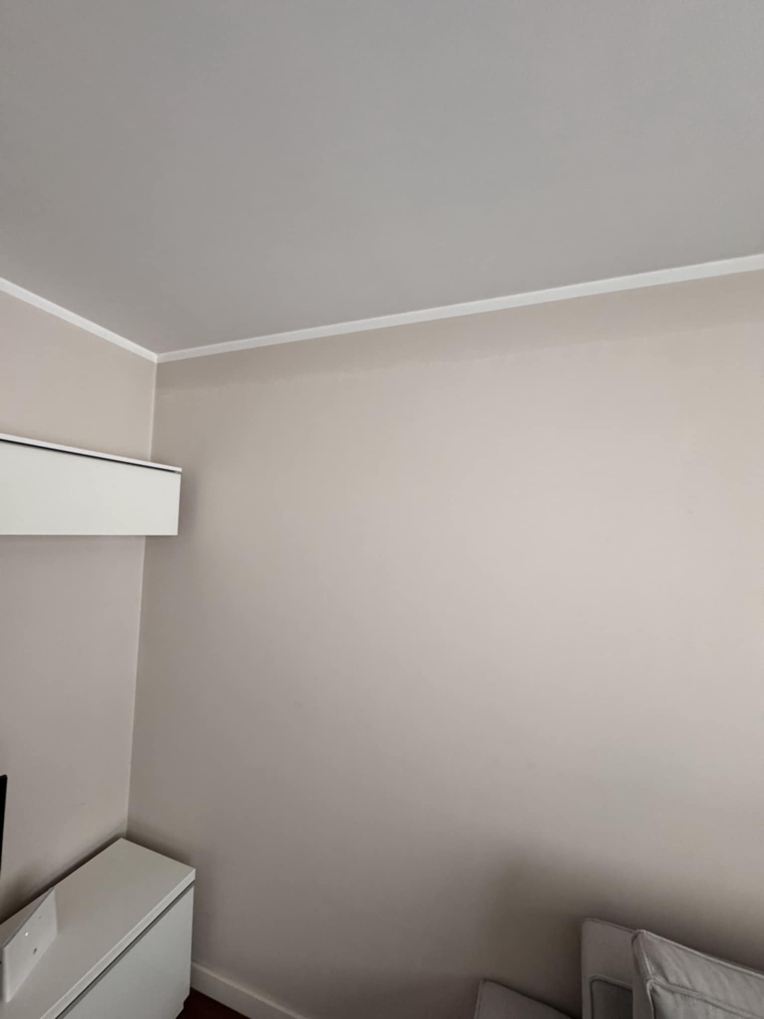 Ściana w odcieniach beżu po malowaniu, z białą listwą przysufitową i klimatyzatorem. Minimalistyczne wnętrze z elementami mebli w dolnej części kadru.