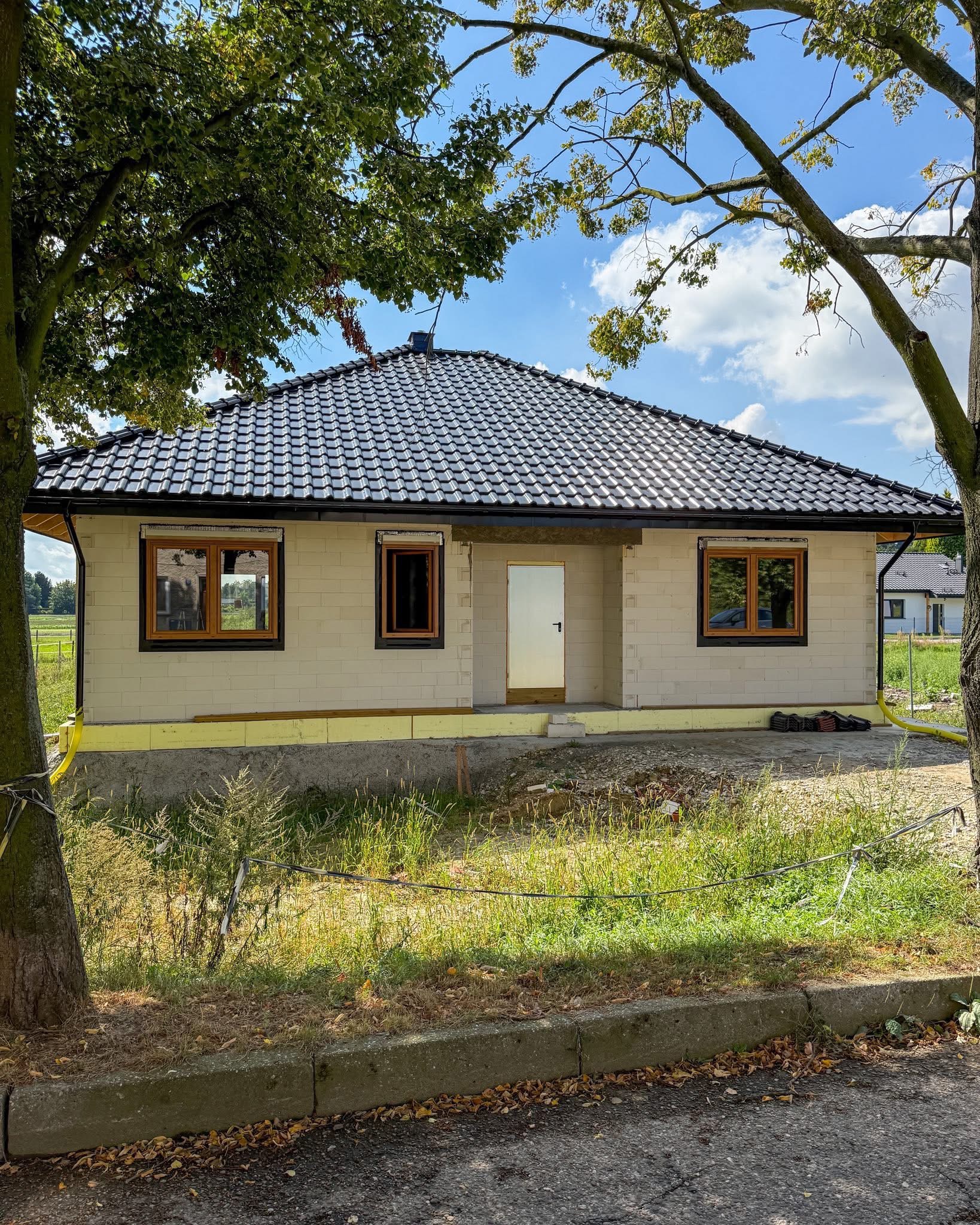 Dom w stanie surowym zamkniętym z oknami z brązowymi ramami w Raciborzu. Widoczne ocieplenie fundamentów i czarna dachówka. Ujęcie z poziomu ulicy.