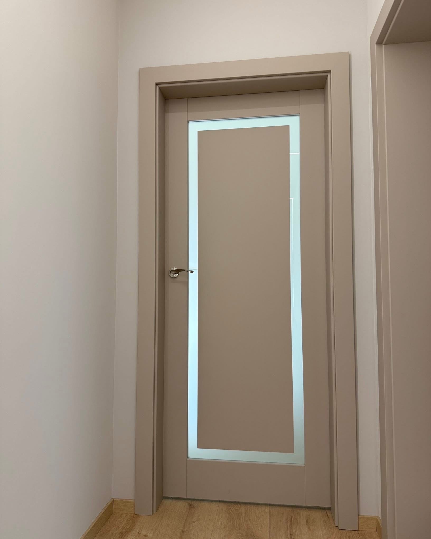 Beżowe drzwi wewnętrzne z prostokątną szybą, srebrna klamka, dopasowana kolorystycznie ościeżnica, drewniana podłoga, jasne ściany. Minimalistyczny design.
