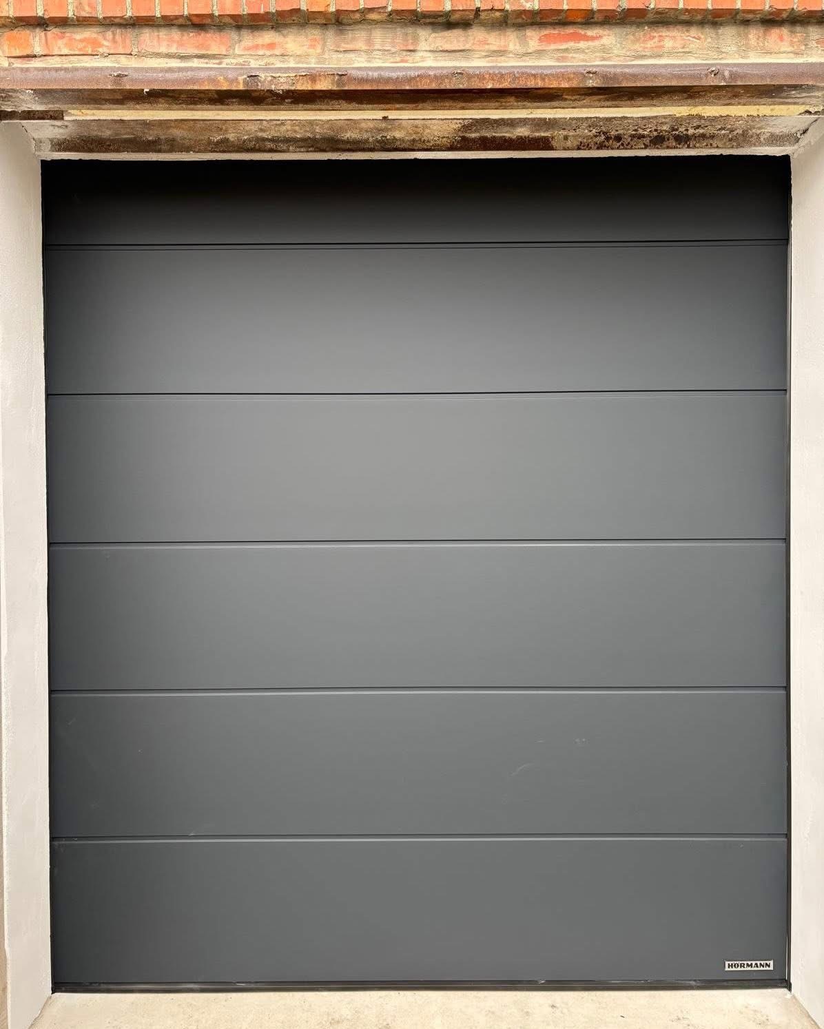 Zamknięta, segmentowa brama garażowa w odcieniach szarości, marki Hormann, z widoczną fakturą paneli i subtelnym oświetleniem, wkomponowana w białą ramę i ceglane nadproże.