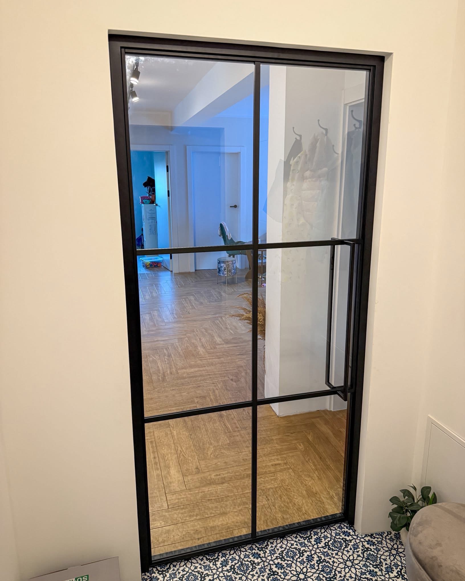 Nowoczesne drzwi szklane z czarną ramą i podziałami, widok na wnętrze z parkietem w jodełkę i wzorzystą podłogą. Minimalistyczny design, jasne ściany.