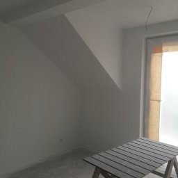 Vladyslav Zabrodin - Poddasze po szpachlowaniu i malowaniu na biało, widoczny stół malarski, okno zabezpieczone folią malarską, surowy beton na podłodze, gniazdko elektryczne.