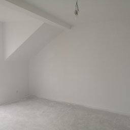 Vladyslav Zabrodin - Wnętrze pokoju po malowaniu na biało, widoczne gładkie ściany i sufit, surowa podłoga, okno zabezpieczone folią, żarówka na kablu. Skos na suficie.