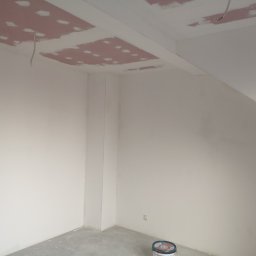 Vladyslav Zabrodin - Wnętrze w trakcie malowania: białe ściany, sufit z widocznymi łatami szpachli, puszka farby w rogu. Widoczne kable elektryczne. Ujęcie z dołu, szeroki kąt.