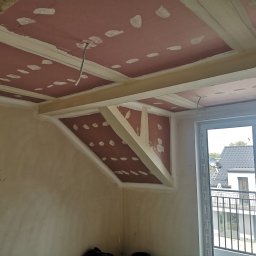 Vladyslav Zabrodin - Poddasze w trakcie adaptacji: widoczne płyty kartonowo-gipsowe na suficie, wzmocnione drewnianymi belkami, przygotowane do szpachlowania. Surowe ściany i okno balkonowe.