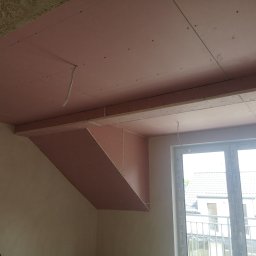 Vladyslav Zabrodin - Montaż płyt karton-gips na suficie poddasza, widoczne profile i łączenia. Przewody elektryczne wystają. Okno z widokiem na domy w tle.