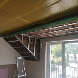 Vladyslav Zabrodin - Skośny sufit z izolacją z wełny mineralnej i metalową konstrukcją, widok z dołu, drabina w rogu, okno balkonowe z widokiem na zewnątrz.