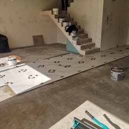 Vladyslav Zabrodin - Układanie białych płytek podłogowych z marmurowym wzorem, z użyciem żółtych krzyżyków dystansowych. W tle schody i puszki z farbą.