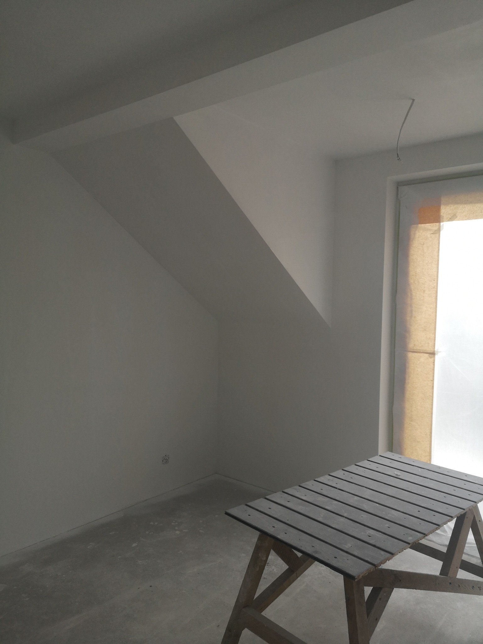 Poddasze po szpachlowaniu i malowaniu na biało, widoczny stół malarski, okno zabezpieczone folią malarską, surowy beton na podłodze, gniazdko elektryczne.