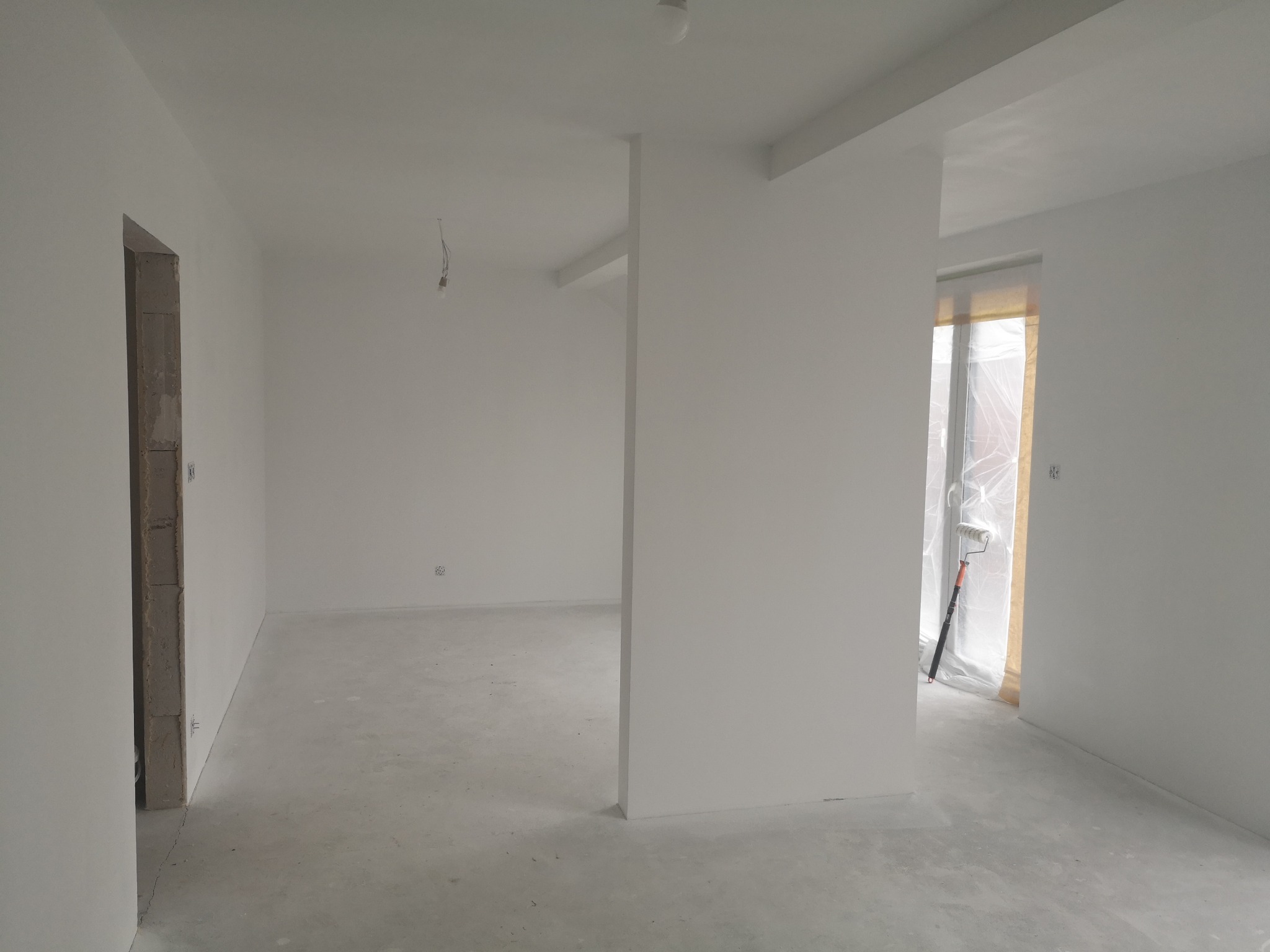Białe, świeżo pomalowane wnętrze z widocznym wałkiem malarskim przy oknie. Surowy beton na podłodze, przygotowane do dalszych prac wykończeniowych.