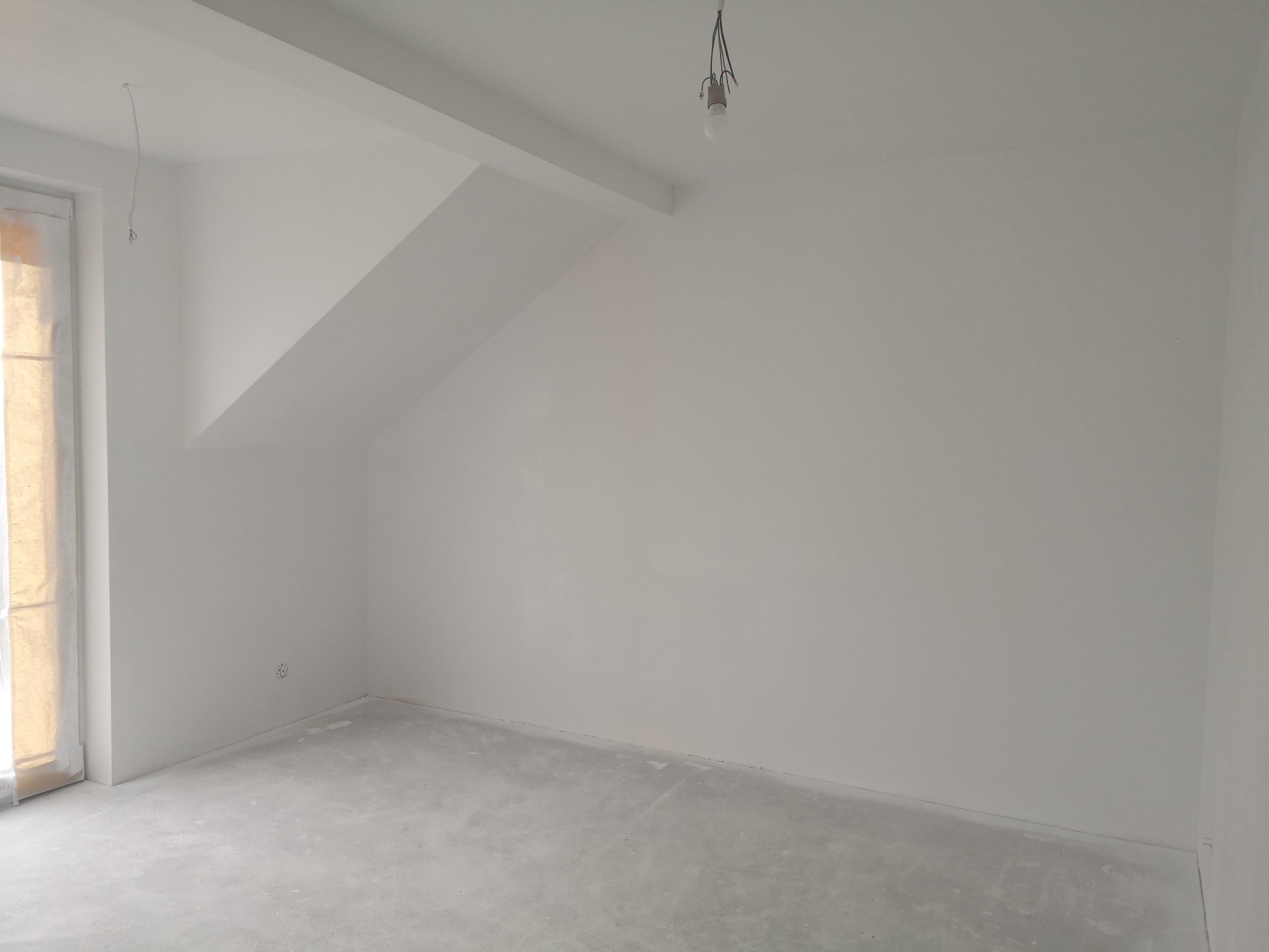 Wnętrze pokoju po malowaniu na biało, widoczne gładkie ściany i sufit, surowa podłoga, okno zabezpieczone folią, żarówka na kablu. Skos na suficie.