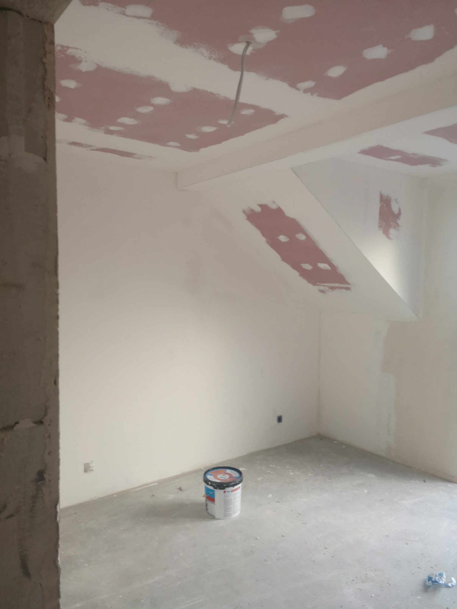 Wnętrze w trakcie prac wykończeniowych: ściany i sufit po szpachlowaniu, widoczne łączenia płyt gipsowych, na podłodze stoi wiadro z farbą. Surowy stan.