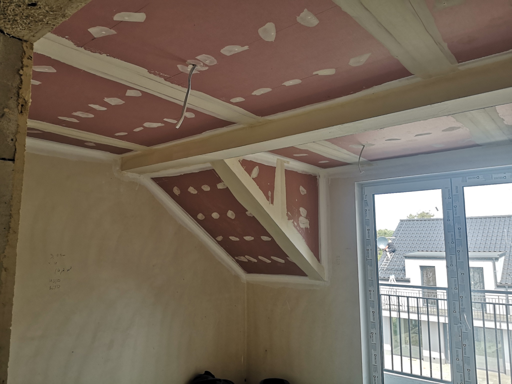 Poddasze w trakcie adaptacji: widoczne płyty kartonowo-gipsowe na suficie, wzmocnione drewnianymi belkami, przygotowane do szpachlowania. Surowe ściany i okno balkonowe.