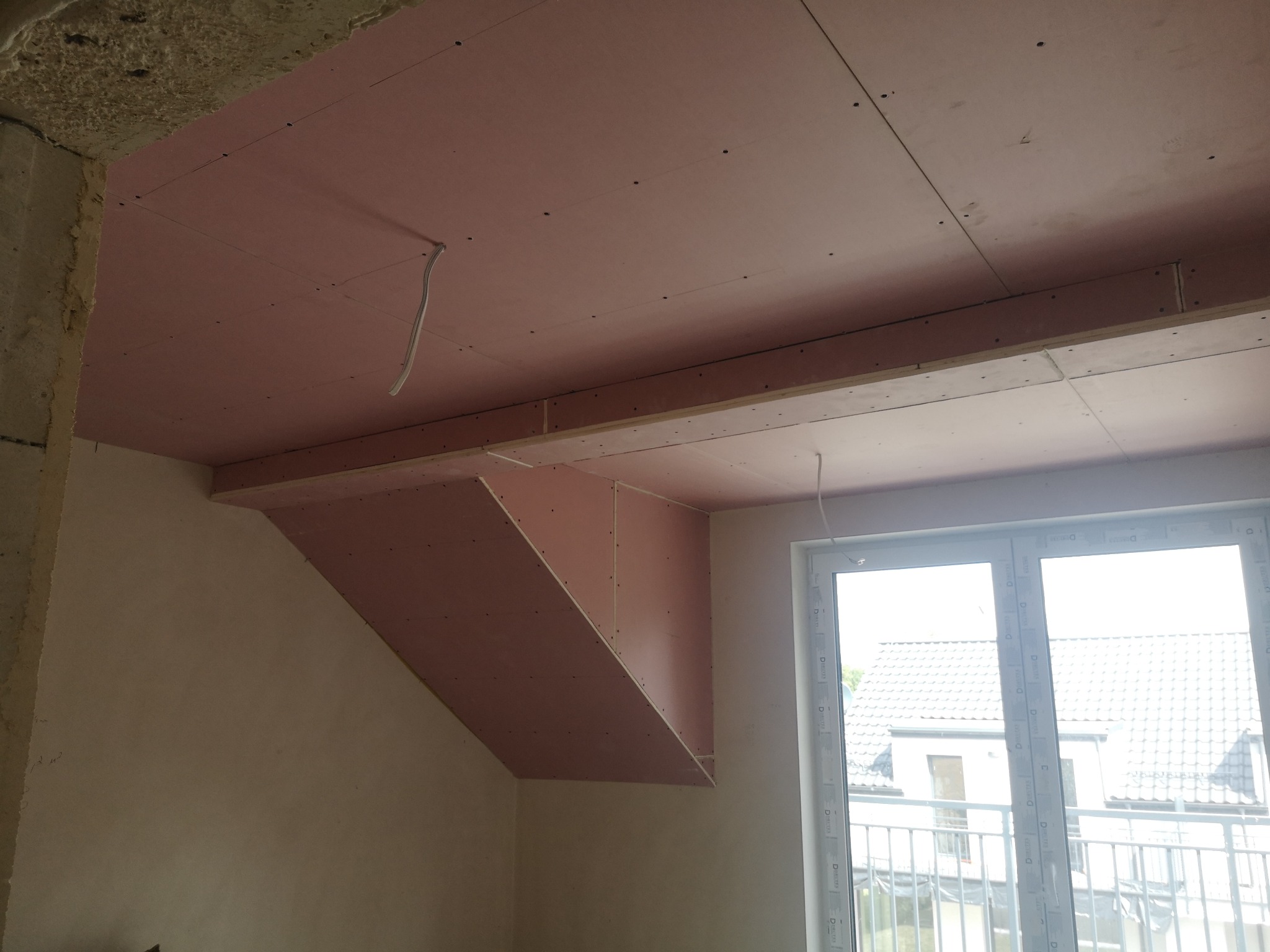 Montaż płyt karton-gips na suficie poddasza, widoczne profile i łączenia. Przewody elektryczne wystają. Okno z widokiem na domy w tle.