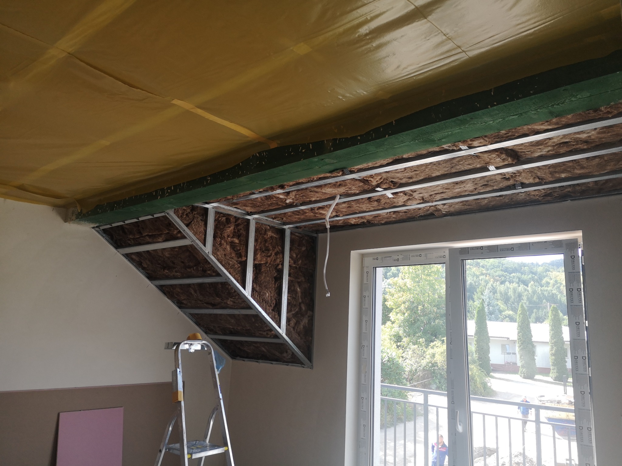 Skośny sufit z izolacją z wełny mineralnej i metalową konstrukcją, widok z dołu, drabina w rogu, okno balkonowe z widokiem na zewnątrz.