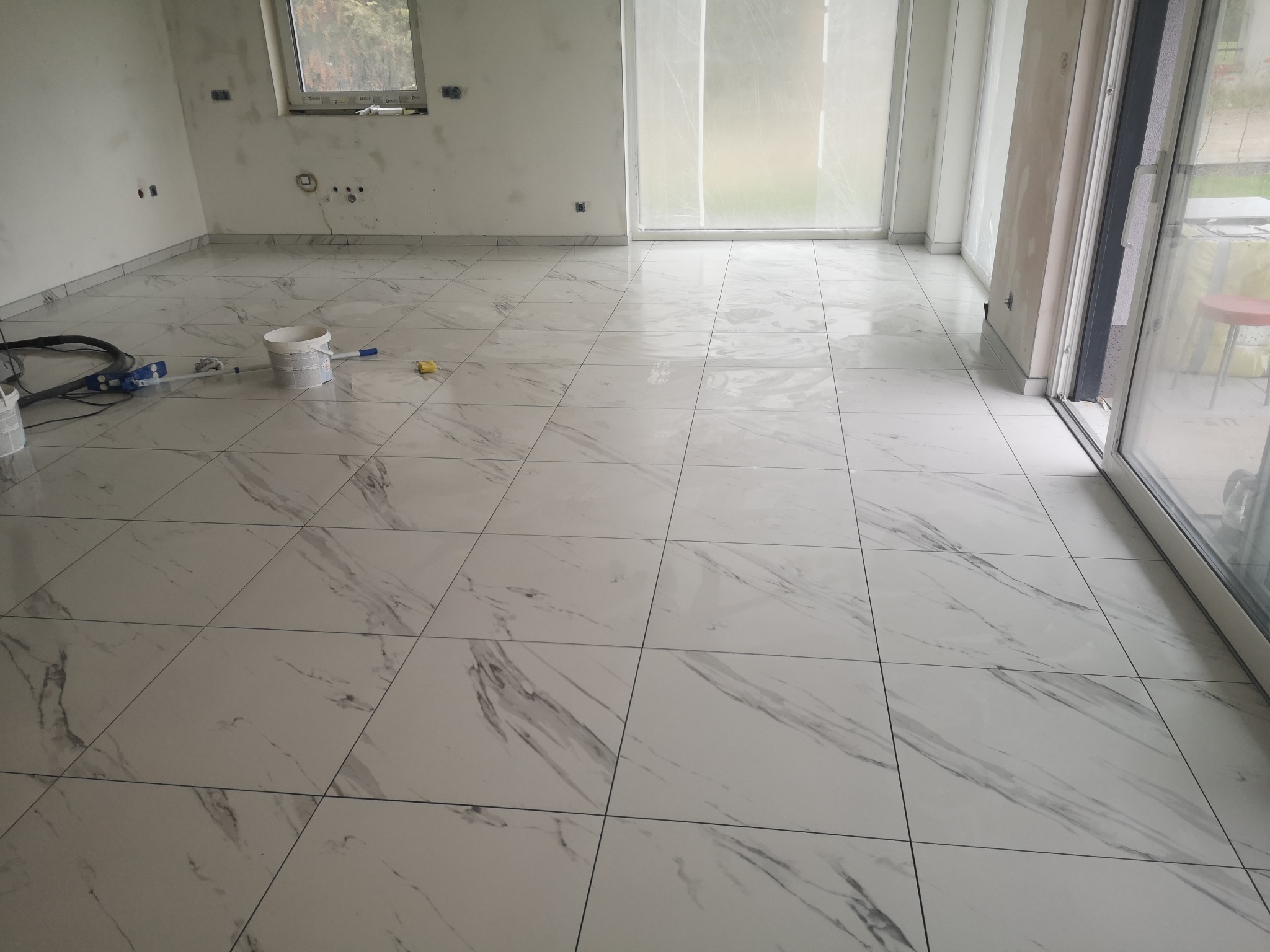 Białe płytki podłogowe imitujące marmur w jasnym, przestronnym wnętrzu. Widoczne narzędzia i akcesoria malarskie, duże okno balkonowe.