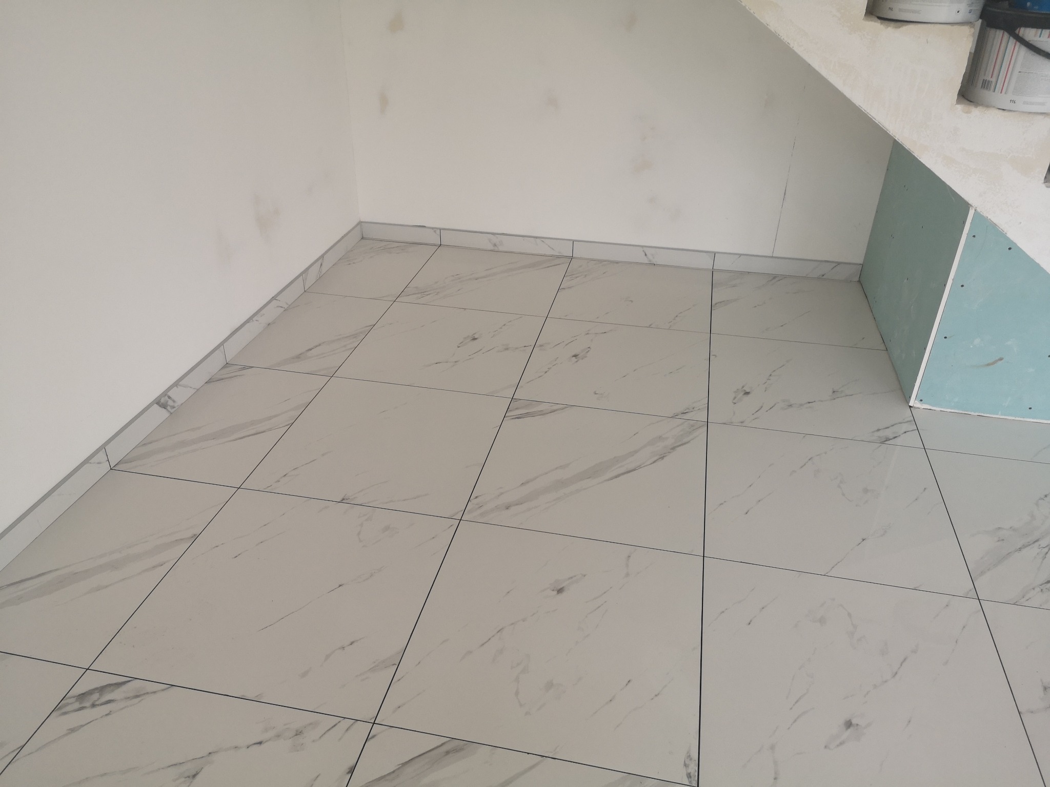 Ukończona podłoga z jasnych, marmurkowych płytek w rogu pomieszczenia. Widoczne białe ściany i fragment płyty karton-gips. Minimalistyczny design, czyste linie.