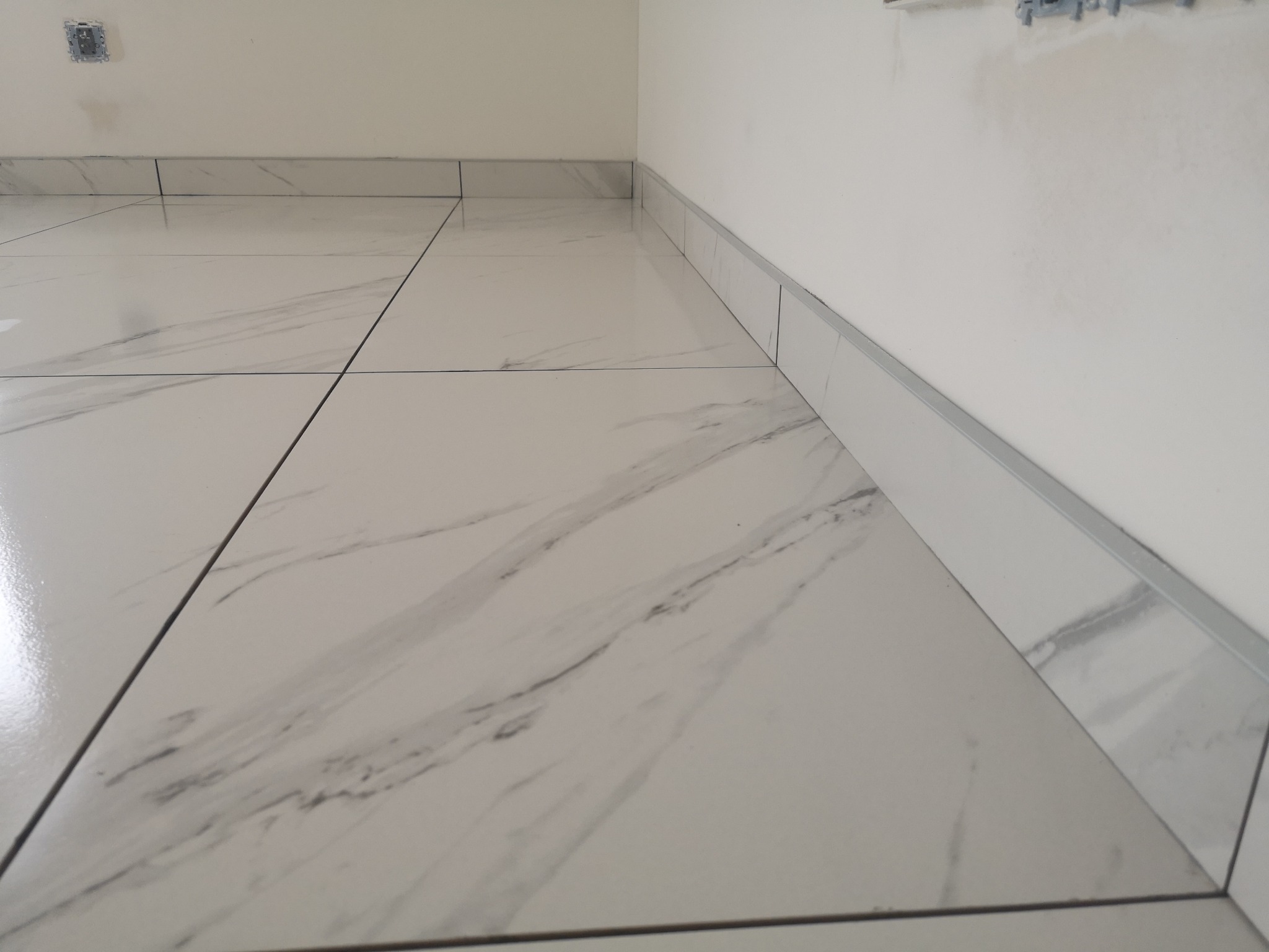 Wykończona podłoga z białych płytek imitujących marmur, z dopasowanymi listwami przypodłogowymi. Widoczny fragment ściany z puszką elektryczną.