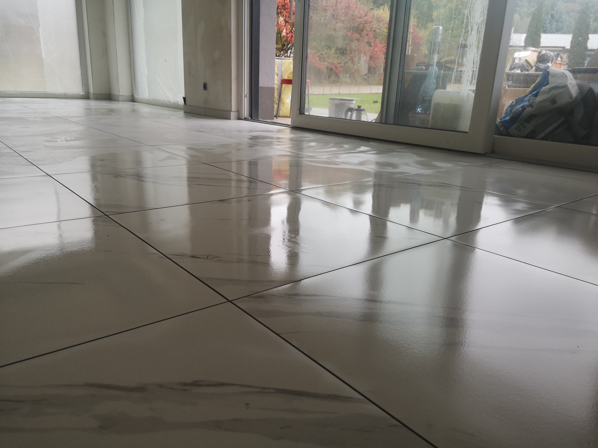 Błyszcząca, jasna podłoga z płytek imitujących marmur, świeżo po ułożeniu. Widoczne odbicia światła i otwarte drzwi balkonowe z widokiem na zewnątrz.