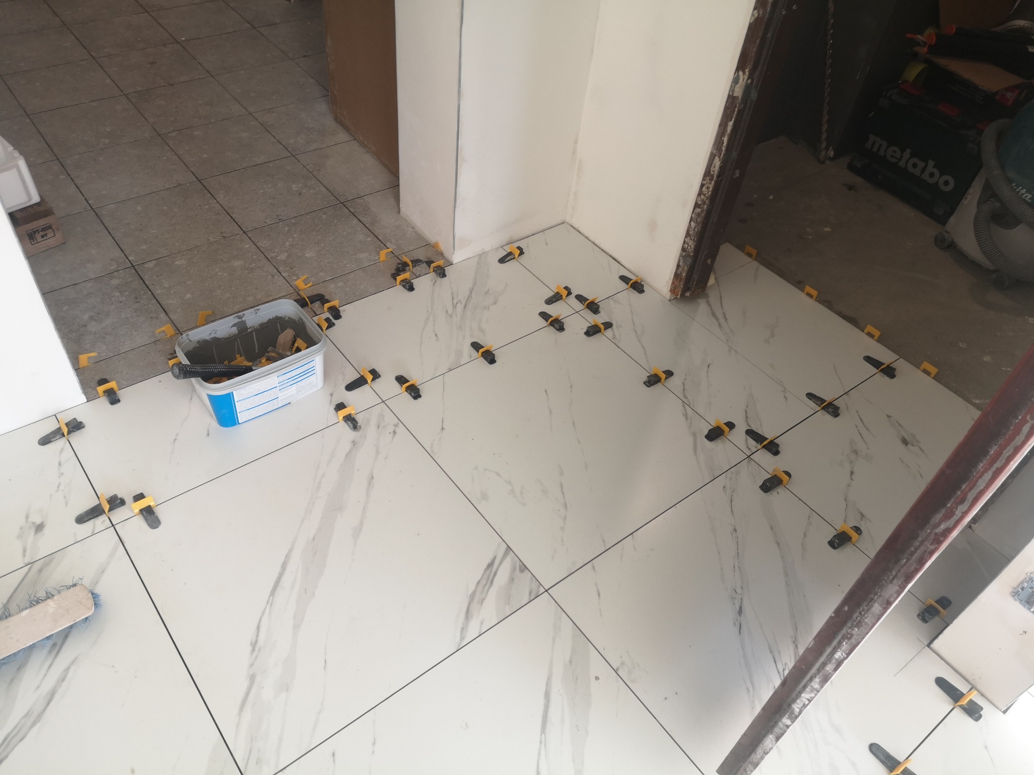 Układanie białych płytek imitujących marmur z użyciem klipsów poziomujących. Widoczny pojemnik z zaprawą i narzędzia. Częściowo ułożona podłoga w trakcie prac wykończeniowych.