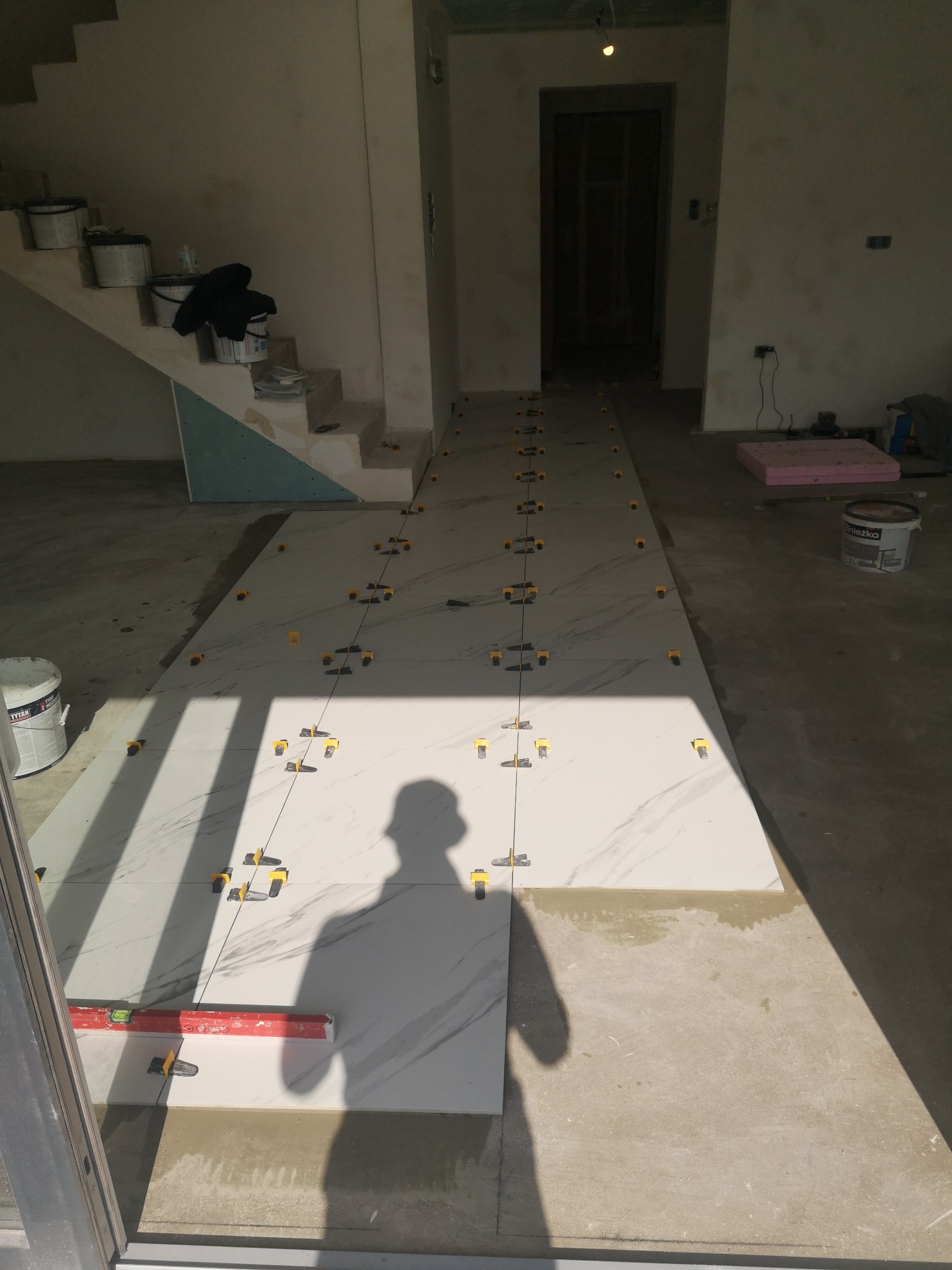 Układanie białych, marmurkowych płytek podłogowych z użyciem poziomicy i klipsów poziomujących. Widoczne schody i puszki z farbą w tle. Jasne światło słoneczne.