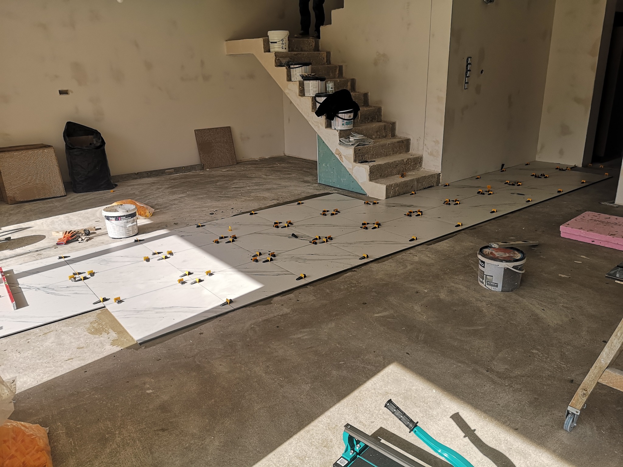 Układanie białych płytek podłogowych z marmurowym wzorem, z użyciem żółtych krzyżyków dystansowych. W tle schody i puszki z farbą.