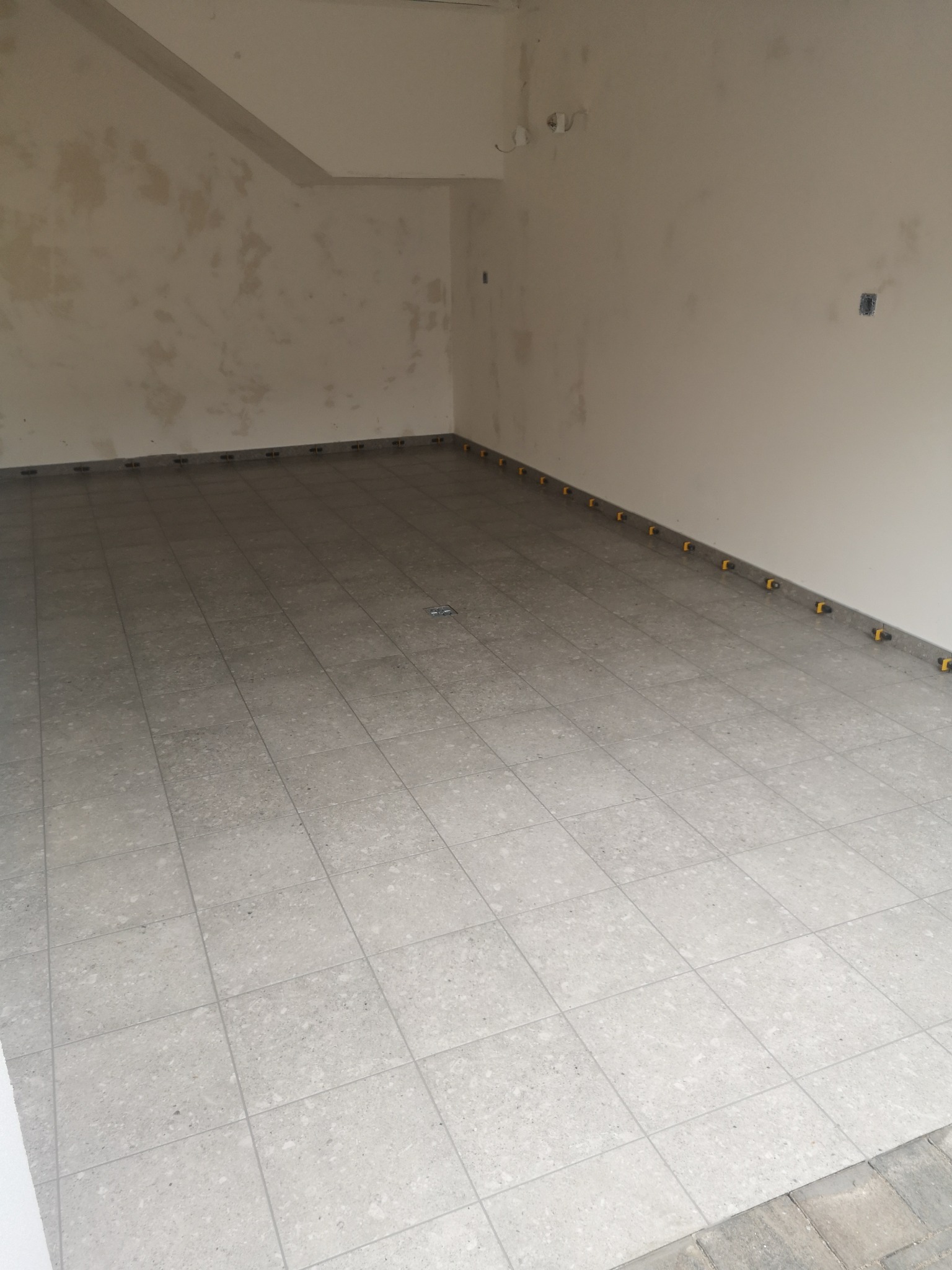 Wykończone płytki podłogowe w jasnym odcieniu szarości, z żółtymi elementami dystansowymi przy ścianach, w surowym wnętrzu przed malowaniem ścian.