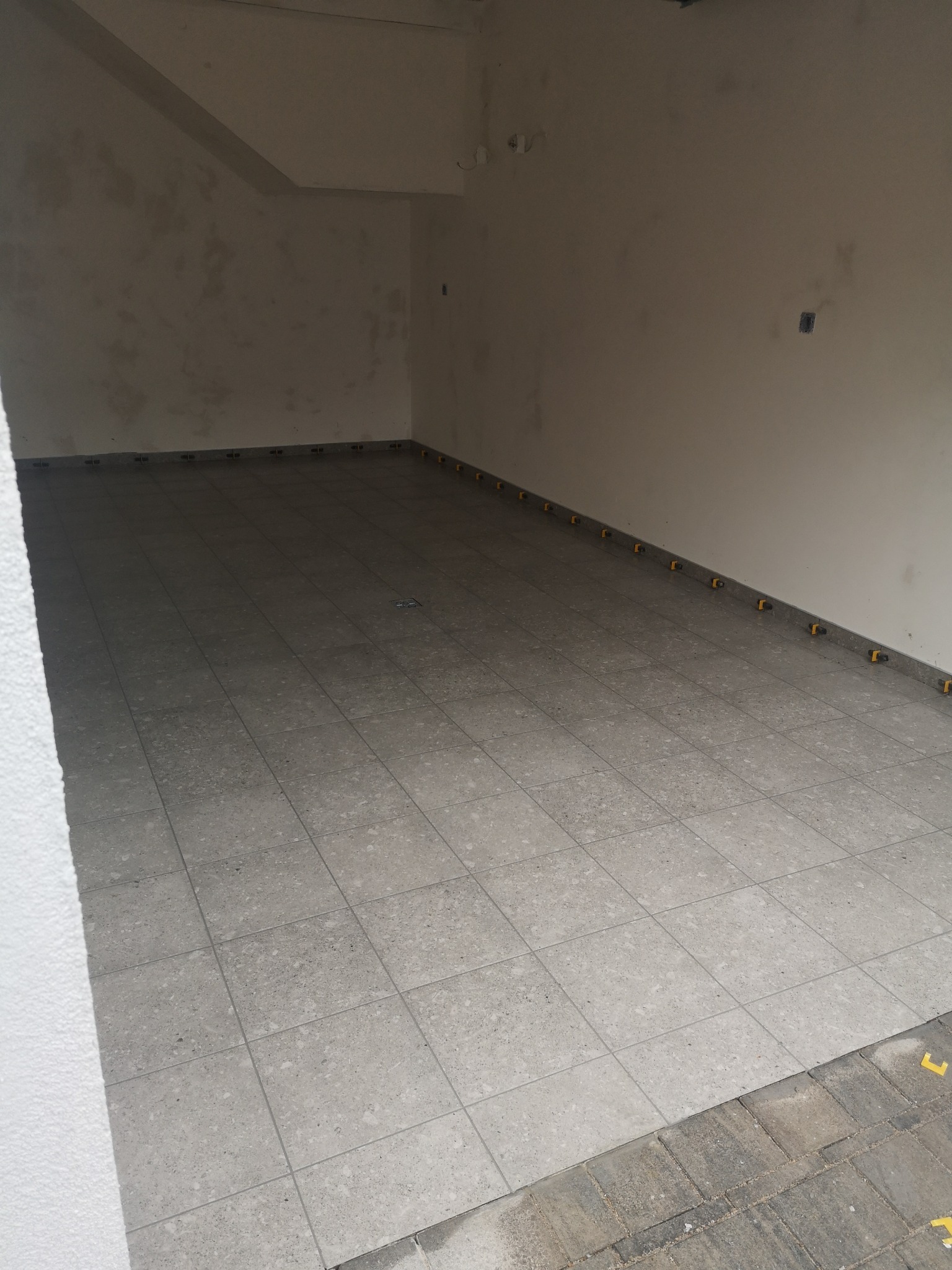 Szare płytki podłogowe ułożone w pomieszczeniu z surowymi ścianami. Żółte kliny dystansowe wzdłuż ściany. Widok z góry, lekki kąt.