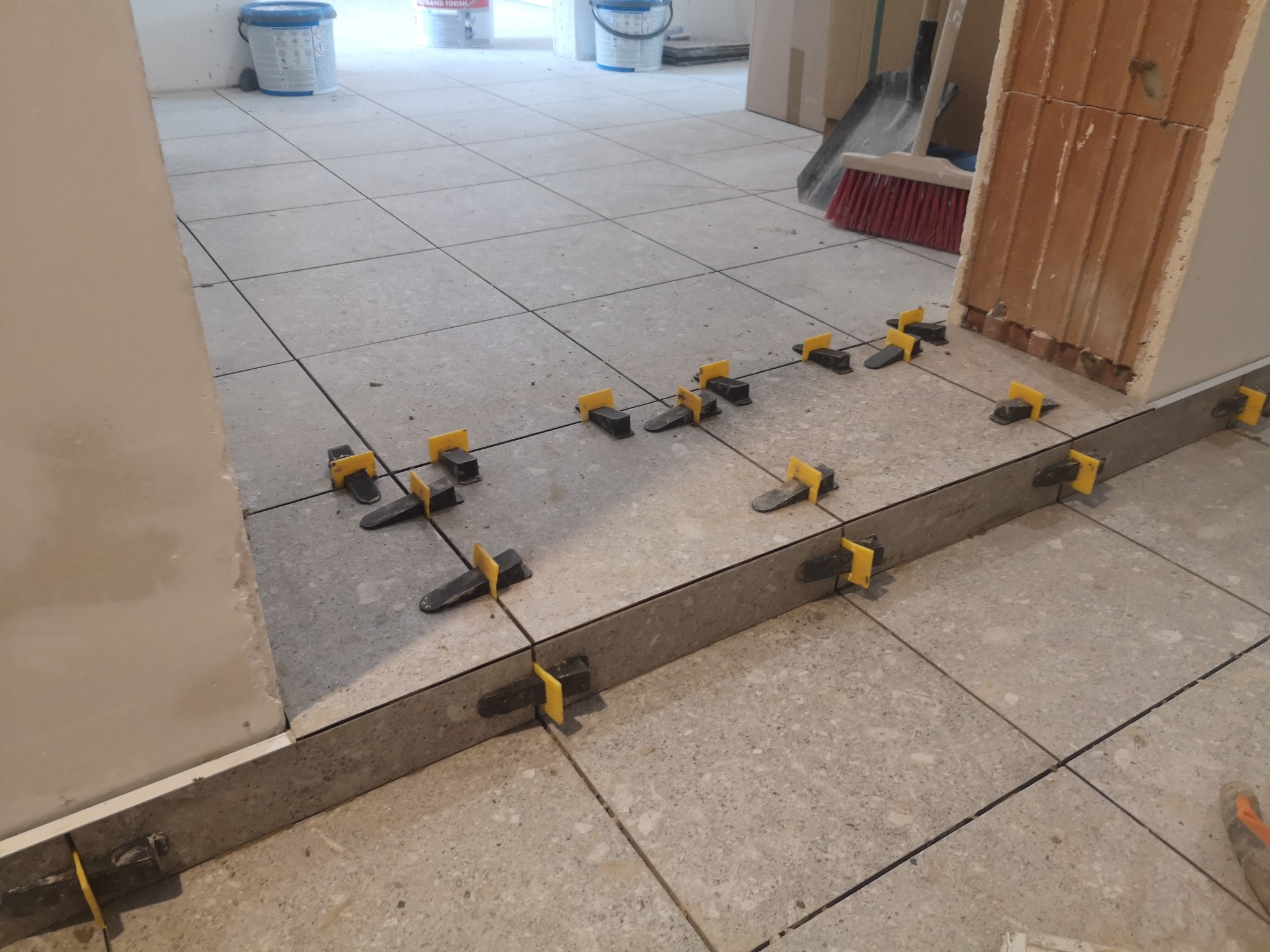 Układanie szarych płytek podłogowych z użyciem systemu poziomowania. Widoczne narzędzia i fragment ściany. Jasne, naturalne światło.