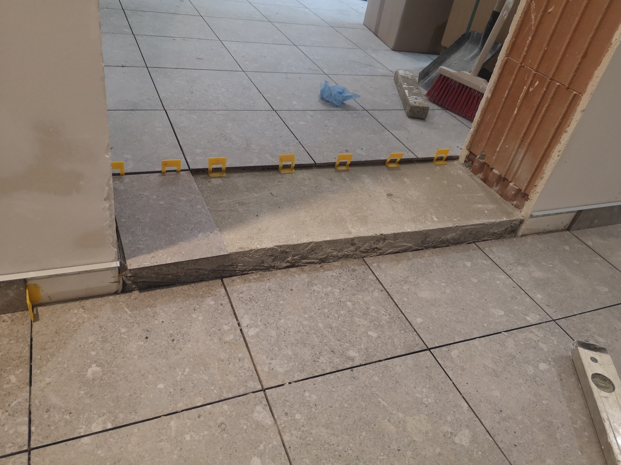 Układanie płytek podłogowych z użyciem żółtych klipsów poziomujących. Widoczny fragment progu i poziomica. Prace w toku, szare płytki.