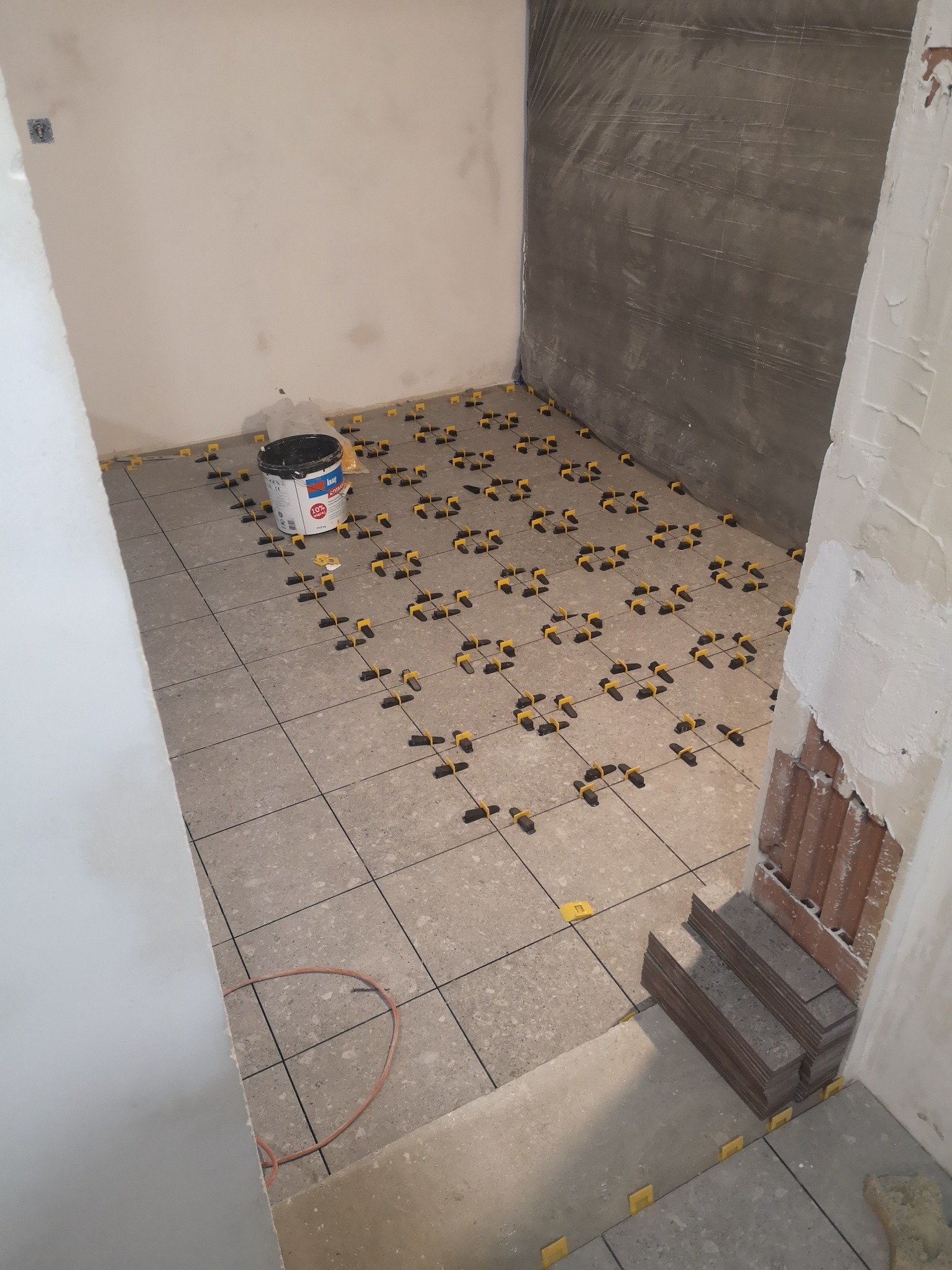 Układanie płytek podłogowych w trakcie prac. Widoczne płytki, system poziomowania, wiadro z klejem i przygotowane płytki do montażu. Ściany w trakcie przygotowania.