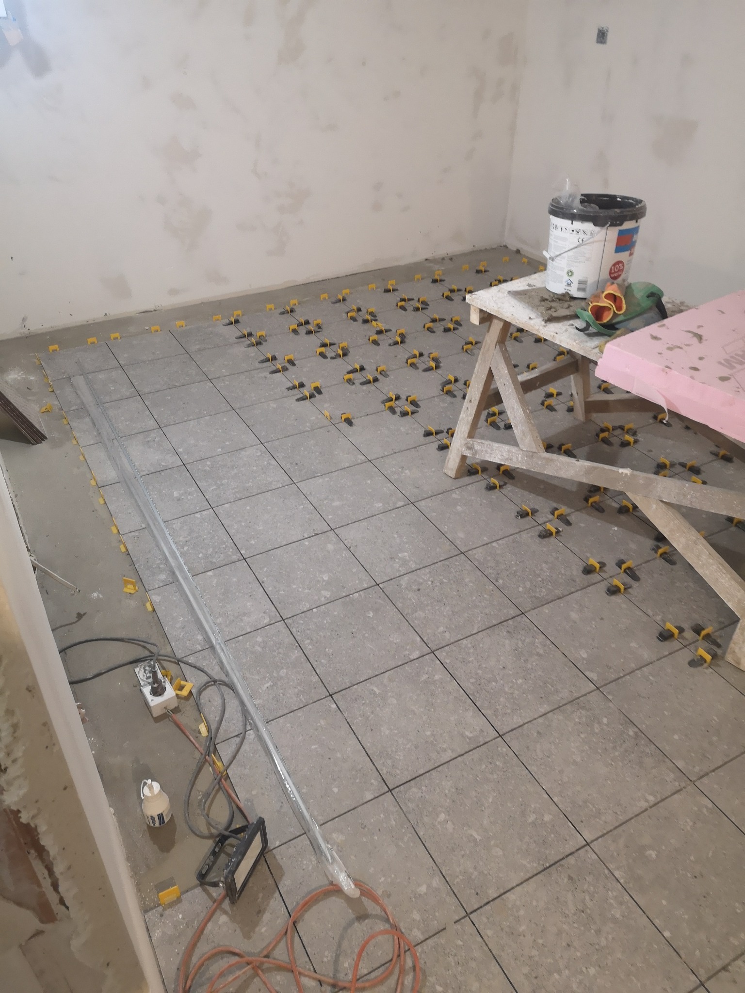 Układanie szarych płytek podłogowych z użyciem żółtych krzyżyków dystansowych. Widoczny stół roboczy z narzędziami i wiadro z klejem. Ściany przygotowane do wykończenia.