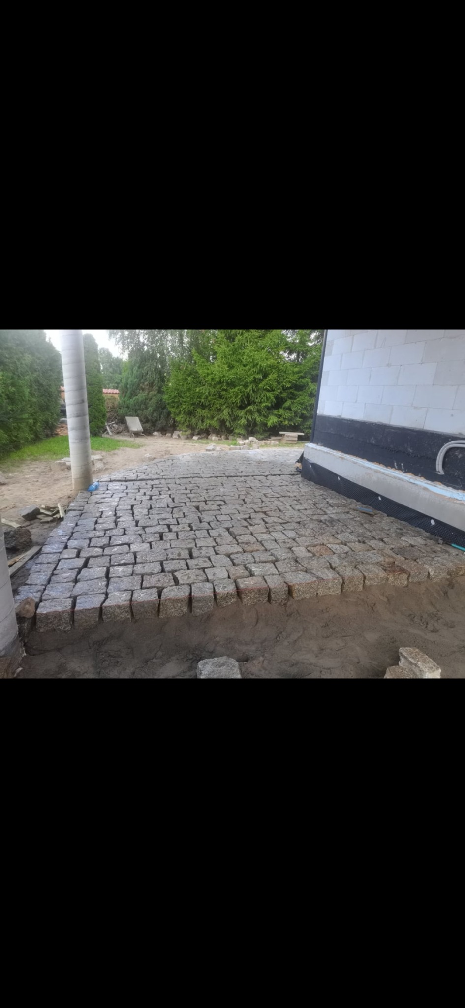 Układanie kostki granitowej przy budynku. Widoczne szare kostki ułożone na podsypce, fragment ściany budynku i zieleń w tle. Prace brukarskie w toku.