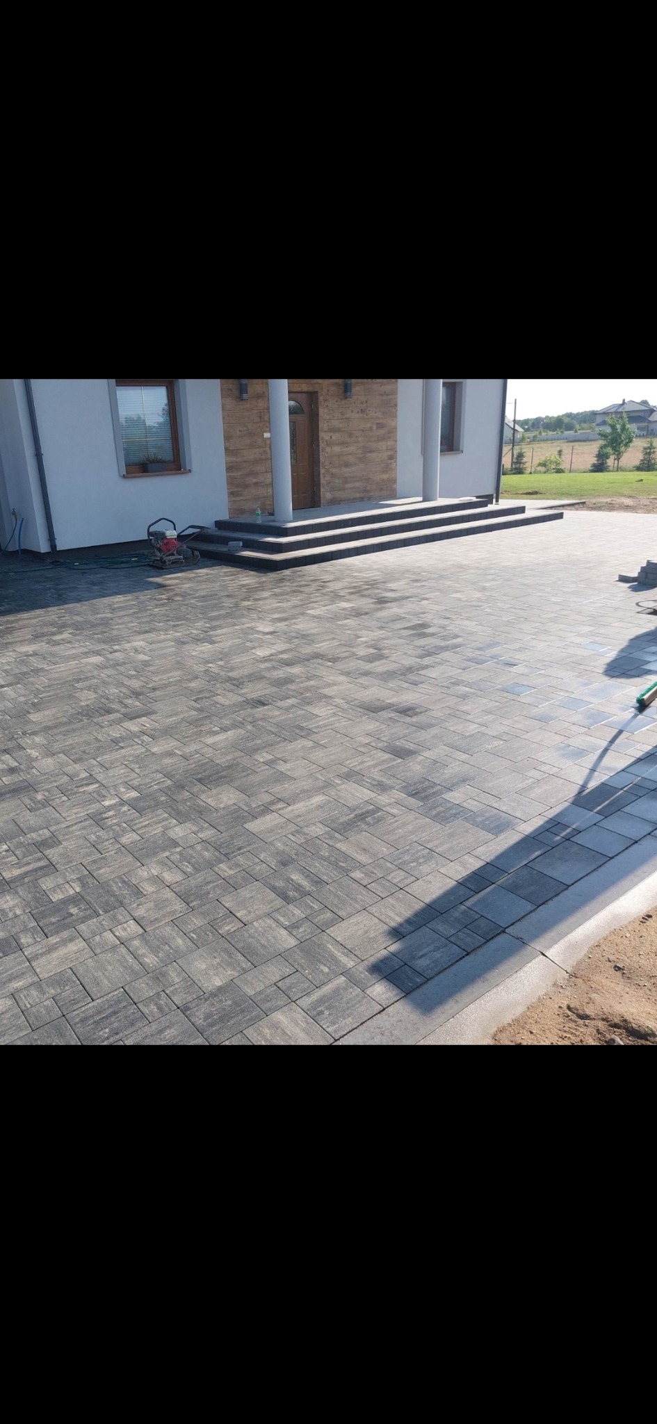 Ułożona kostka brukowa w odcieniach szarości prowadząca do schodów wejściowych domu. Widoczny fragment obrzeża i ubijarka w tle.