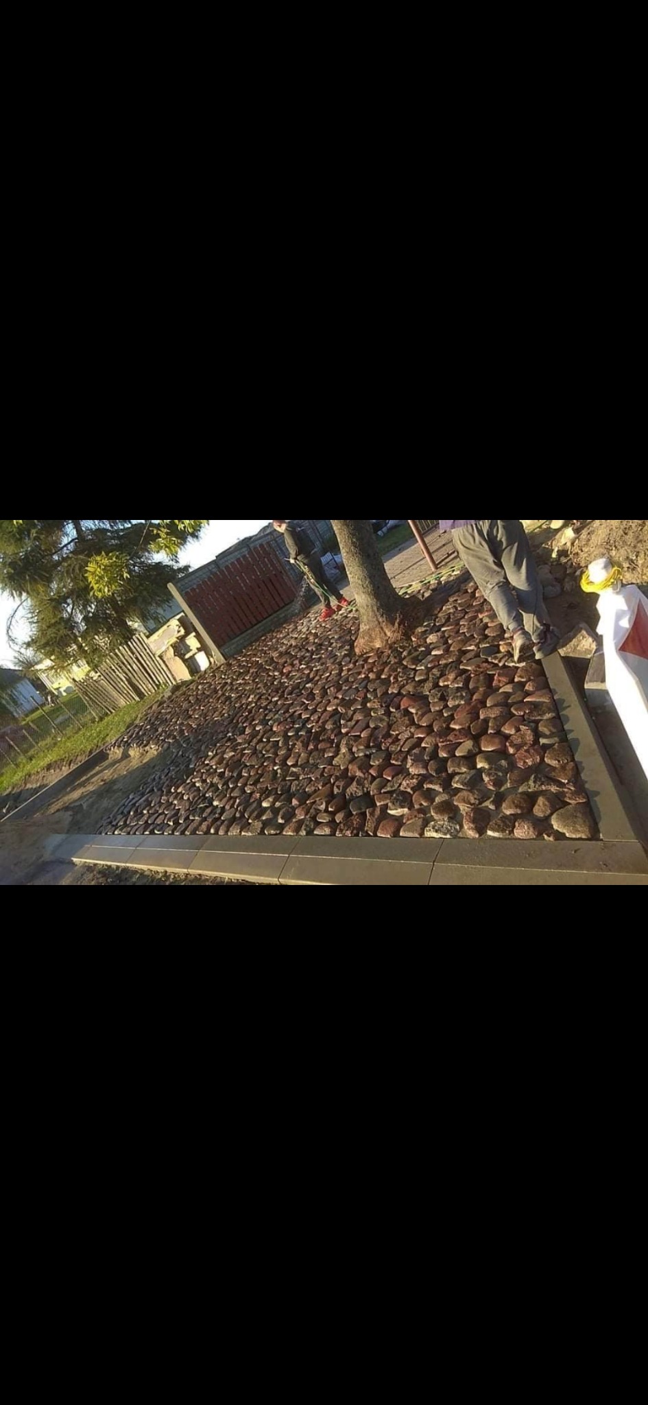 Ścieżka z kamieni polnych obramowana betonową obwódką, otaczająca drzewo. W tle widoczny fragment ogrodzenia i trawnika. Dwóch mężczyzn w tle. Słoneczny dzień.