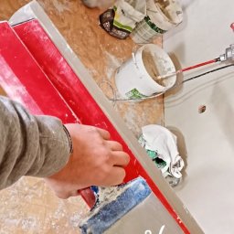 Mateusz 2TUR - Praca z pacą na ścianie, widoczne wiaderka z masą i narzędzia. Przygotowania do nakładania gładzi. Podłoga zabezpieczona przed zabrudzeniem. Ujęcie z góry.