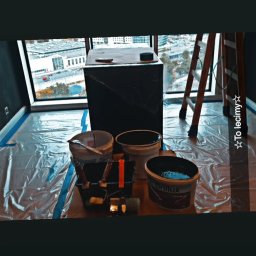 Mateusz 2TUR - Przygotowanie do malowania: wiaderka z farbą Brillux, kuweta z wałkami, drabina, folia ochronna na podłodze, widok na miasto z okna. Etap przed malowaniem ścian.