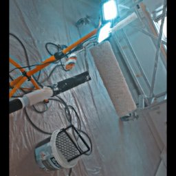 Mateusz 2TUR - Przygotowanie do malowania ścian: wałek, farba, oświetlenie na statywie i folia ochronna. Widok z dołu, podkreślający profesjonalne wyposażenie.