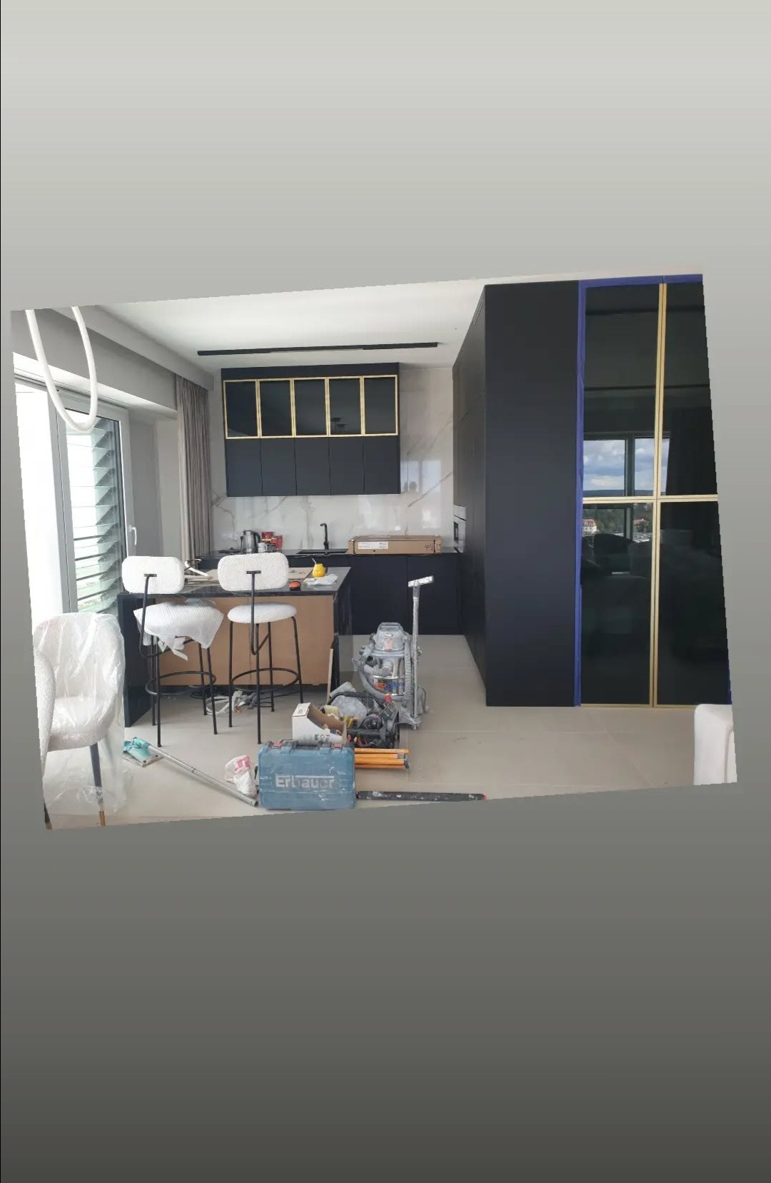 Nowoczesna kuchnia w trakcie wykańczania: czarne szafki z złotymi akcentami, sprzęt budowlany, krzesła barowe. Widok na okno i elementy wykończeniowe.