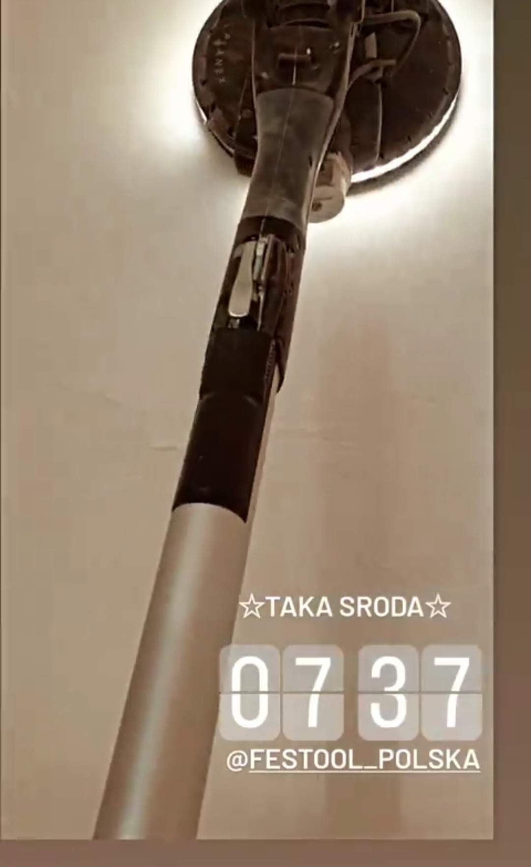 Szlifierka do gładzi marki Festool w trakcie pracy na jasnej ścianie. Widoczny pył i ślady szlifowania. Ujęcie z dołu, narzędzie skierowane ku górze.