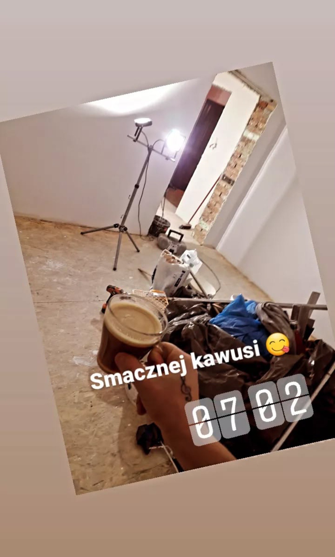 Wnętrze w trakcie prac z lampą na statywie, widoczne narzędzia i kubek z kawą w dłoni. Fragment ściany z odsłoniętą cegłą. Ujęcie z bliska, z lekkim obrotem.