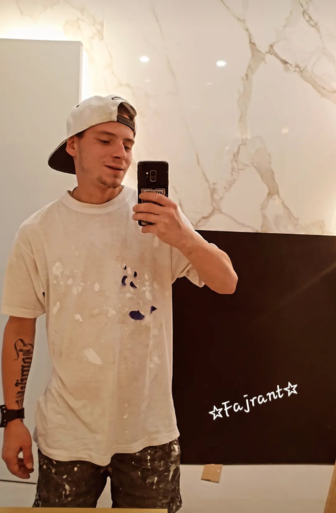 Malarz w ubrudzonym farbą t-shircie robi selfie na tle wykończonej ściany z płytek imitujących marmur i czarnej ściany. Widoczne plamy farby na ubraniu.