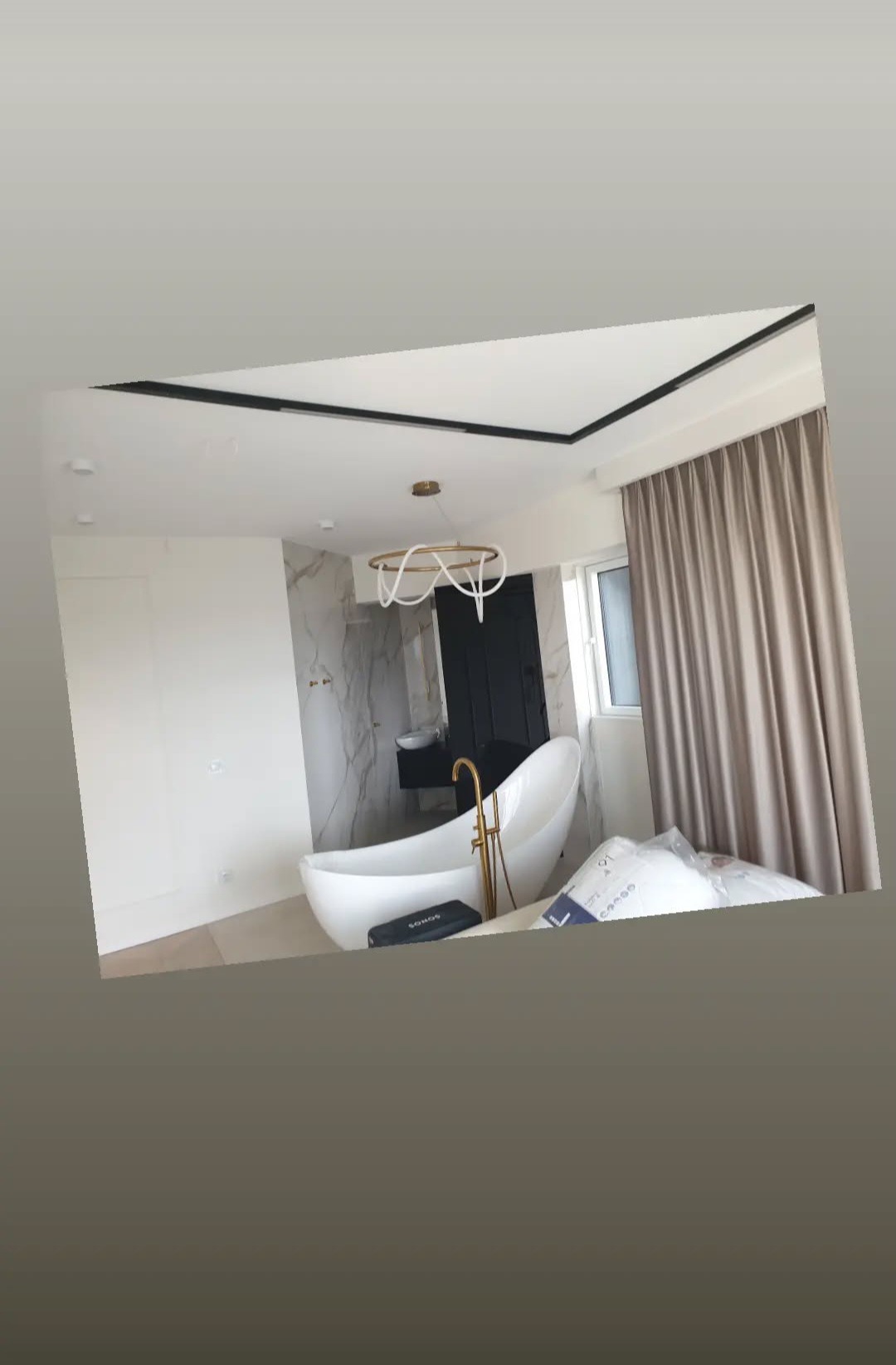 Nowoczesna łazienka z wolnostojącą wanną, złotą armaturą i marmurową ścianą. Czarna listwa sufitowa i beżowe zasłony dodają elegancji. Widoczny głośnik Sonos.