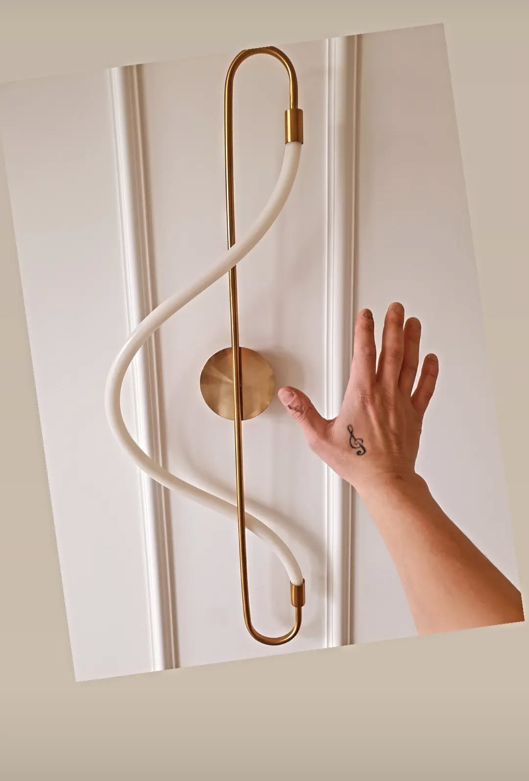 Biała ściana z dekoracyjną lampą w kształcie klucza wiolinowego. Dłoń z tatuażem klucza wiolinowego dotyka lampy. Minimalistyczny design.