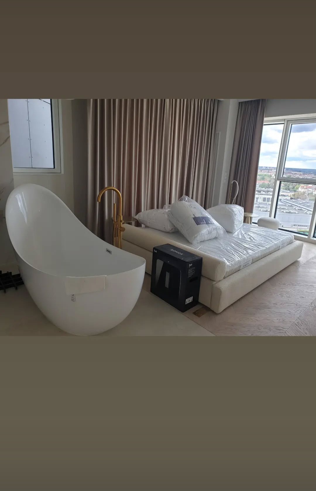Nowoczesna łazienka w apartamencie z wolnostojącą wanną i złotą armaturą. W tle łóżko z pościelą w folii i widok na miasto przez okno.