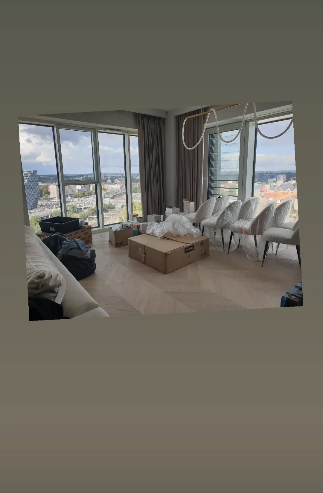 Wnętrze apartamentu w trakcie urządzania: kartony, meble zabezpieczone folią, widok na miasto przez panoramiczne okna. Parkiet ułożony we wzór jodełki.
